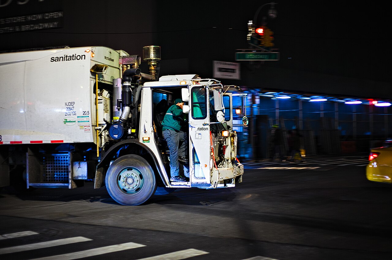 A New York City Department of Sanitation garbage truck turning onto Broadway in the evening