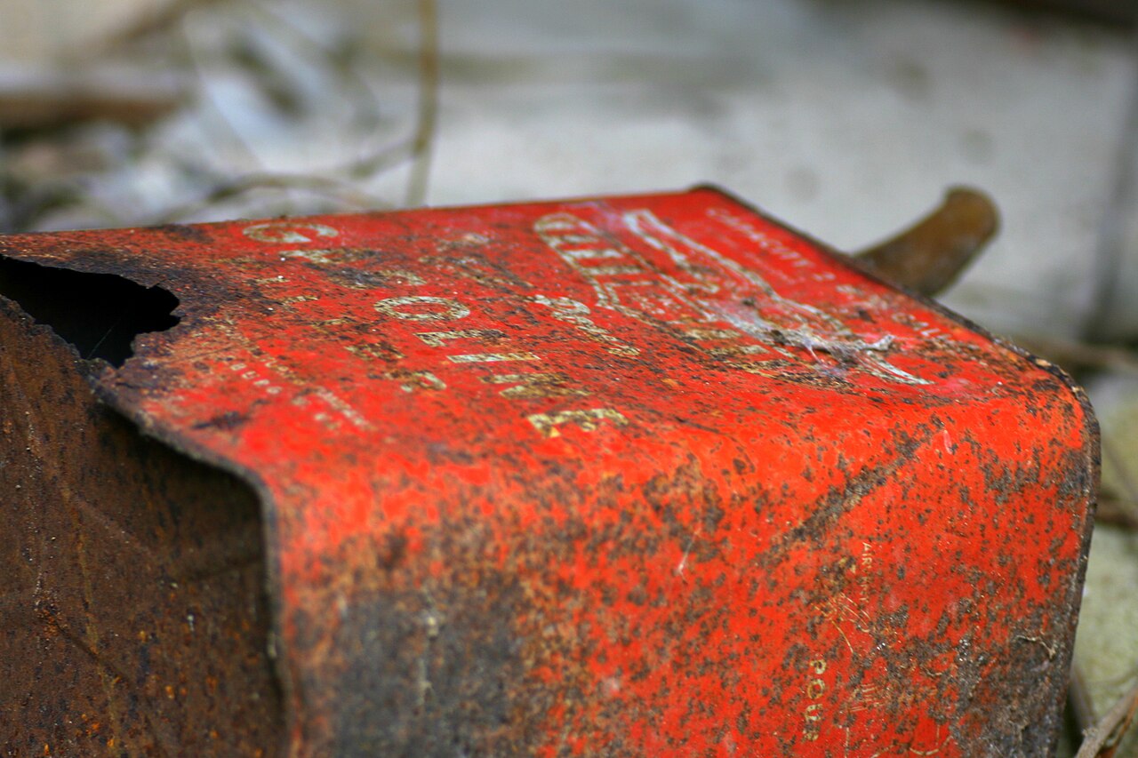 A rusted, dented old red metal gas can lying on the ground