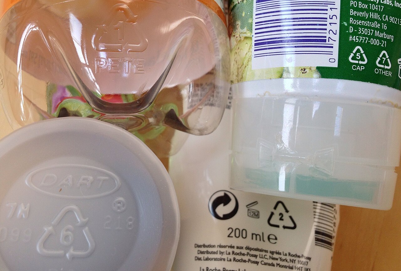 Plastic packaging with molded and printed resin identification codes: 1 PETE on a clear bottle, 6 on a white lid, 2 on a lotion tube, and 5 and 7 on a printed label